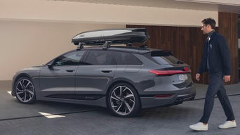 Audi mit einer Dachbox steht vor einem Haus und ein Mann mit Brille läuft aus diesen zu