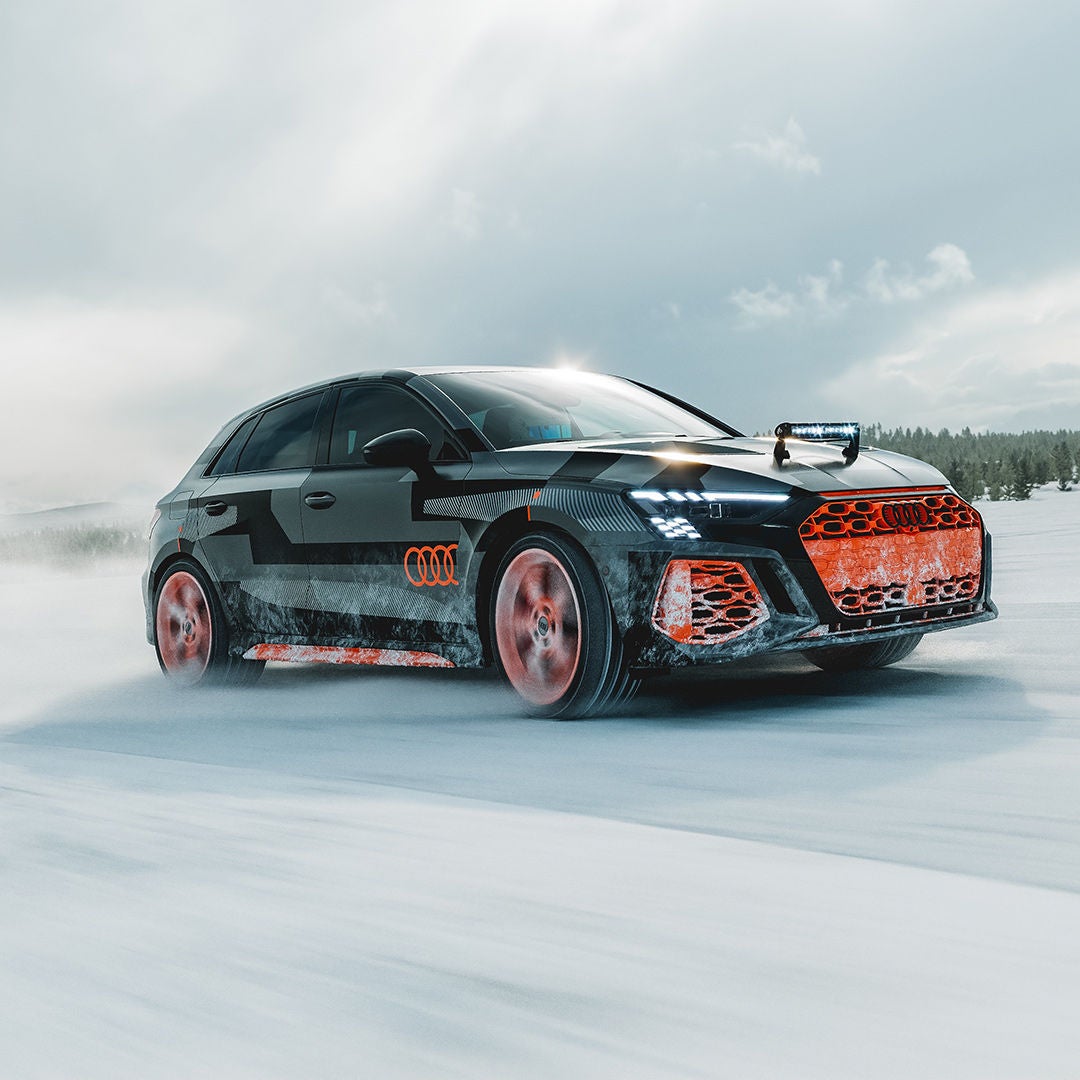 A black Audi RS 3, featuring orange accents, drives across a snow-covered area; snow dust, headlights on, forest in the background.