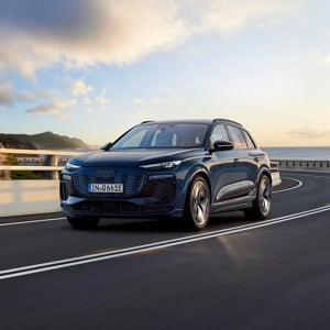 A dark blue Audi SUV drives along a coastal highway at sunset, with a scenic ocean and mountain backdrop. The scene conveys freedom and adventure.