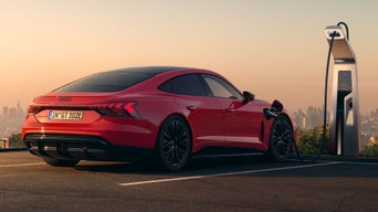 Ein roter Audi e-tron GT quattro steht auf einer Anhöhe über einer Stadt, die ins Licht eines Sonnenuntergangs gehüllt ist. Das Fahrzeug wird an einer öffentlicher Ladesäule geladen.