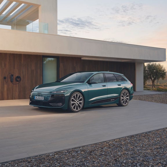 A sleek green electric car parked in front of a modern house with wood paneling. Charging station visible.