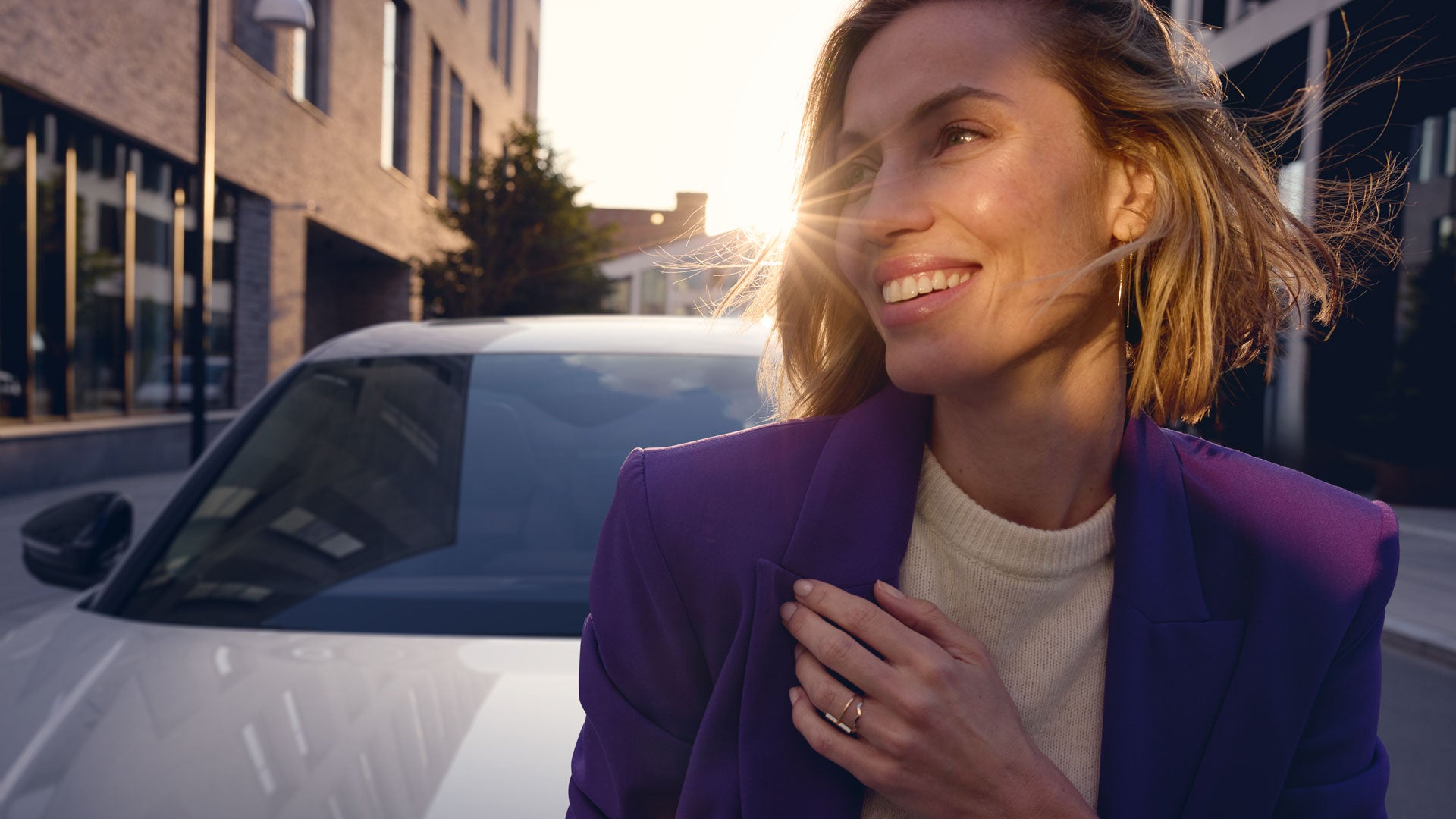 Lächelnde Frau im lilafarbenen Blazer vor einem Auto im Abendlicht.