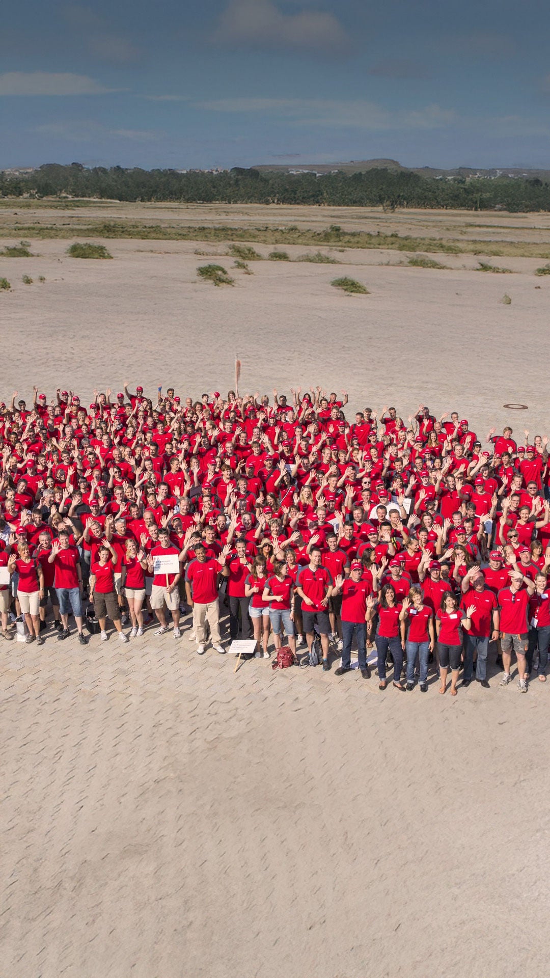 Luftaufnahme einer großen Gruppe von Audi Mitarbeitenden in roten Shirts. Sie stehen vor dem Firmensitz und winken in die Kamera.