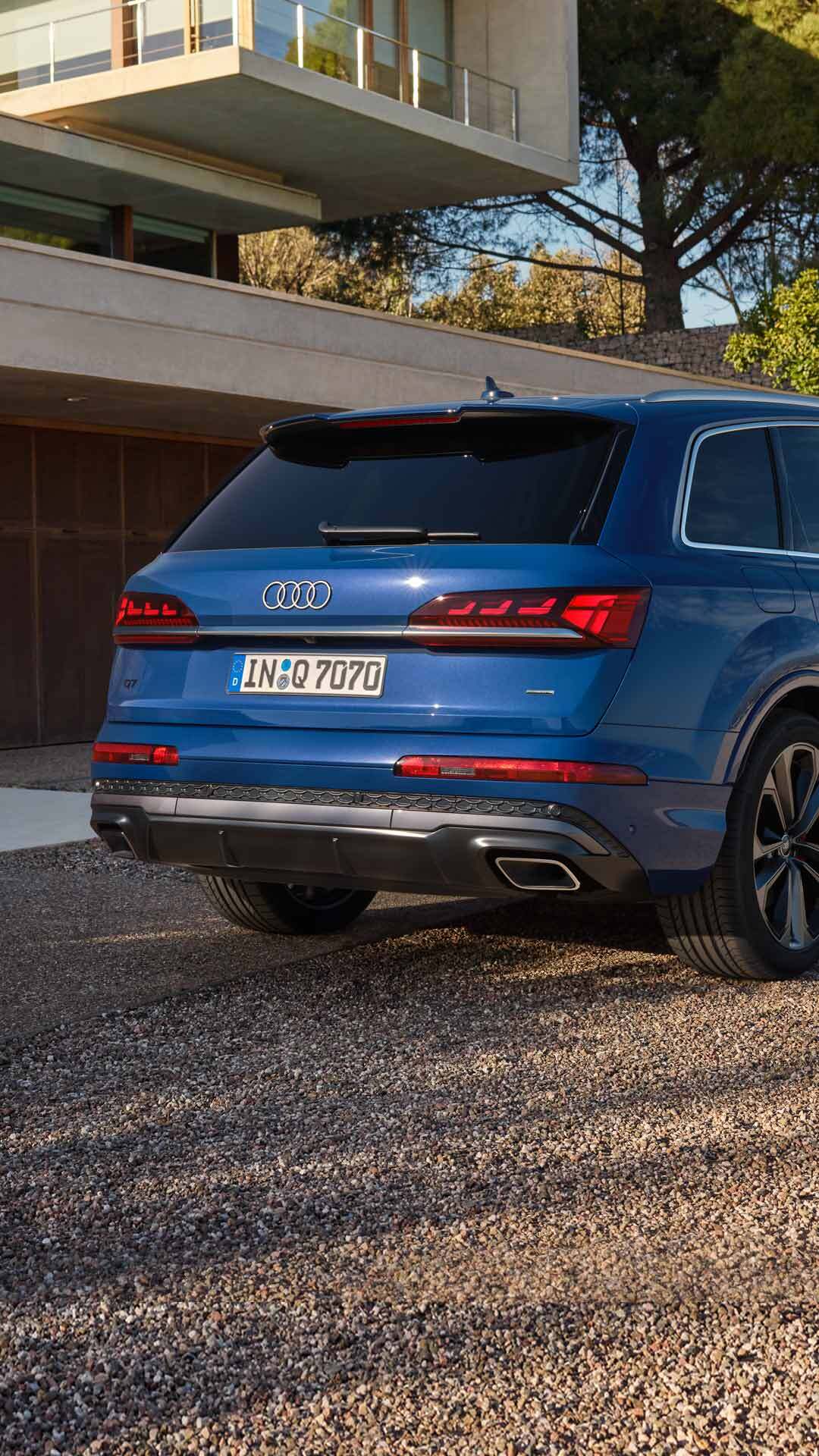 Ein blauer Audi Q7 SUV seitlich vor moderner Fassade, Paar im Businesslook steht lächelnd im Hintergrund.
