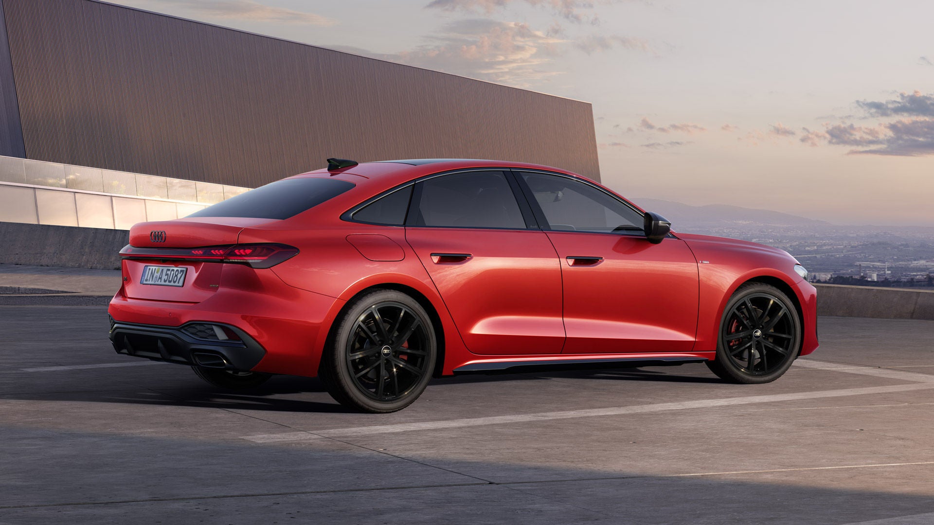 Die A5 Limousine im schrägen Seitenprofil. Das Fahrzeug ist misanorot lackiert und steht auf einem Parkdeck vor einem großen Gebäude.