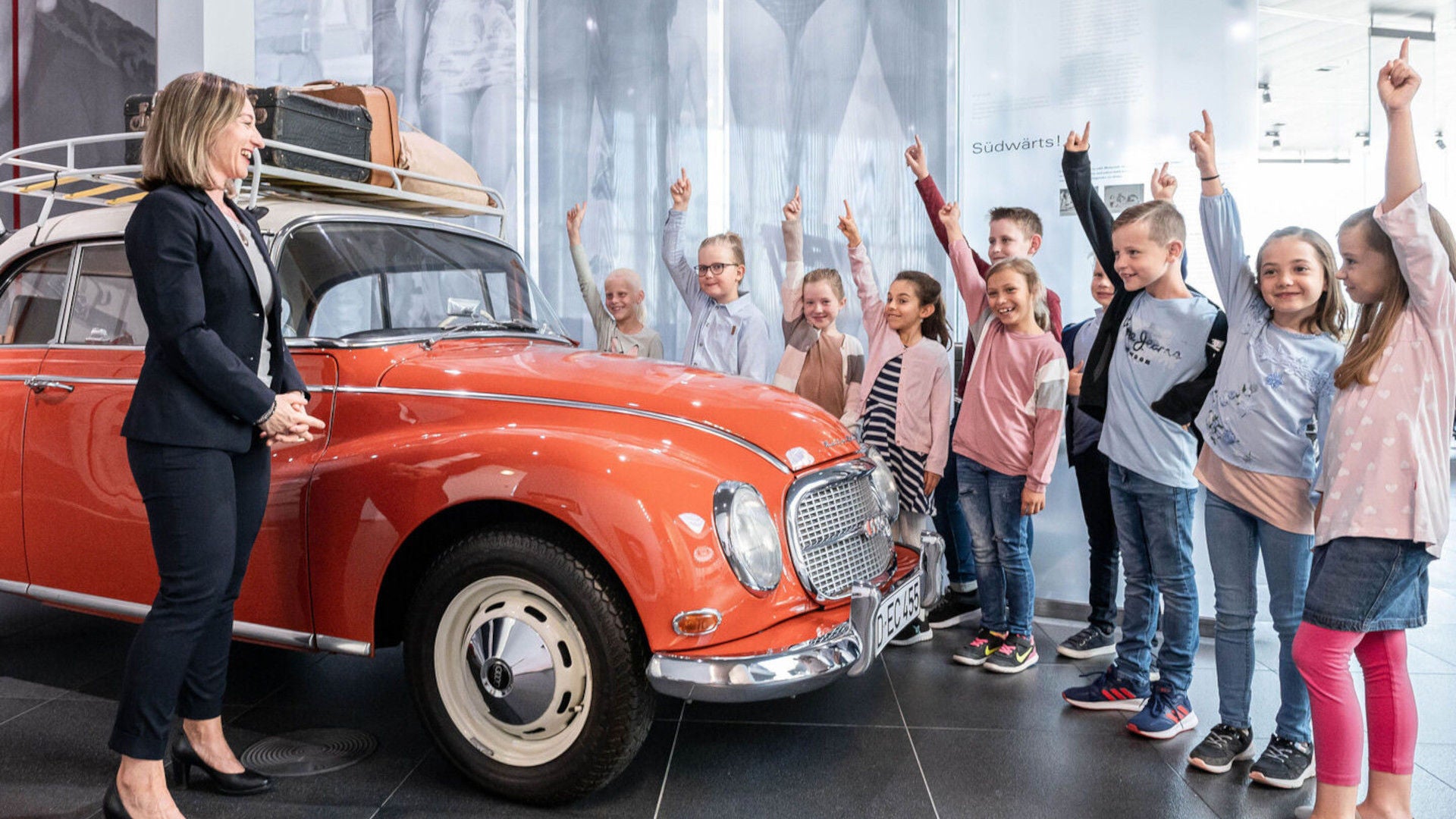 Frau und Schulkinder im Museum neben rotem Oldtimer mit Dachgepäckträger, Kinder zeigen nach oben