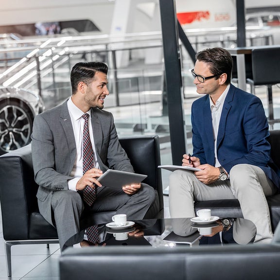 Zwei Männer im Anzug führen Gespräch mit Notizen und Tablets in moderner Lounge mit Kaffeetassen, im Hintergrund ein Audi Modell. 