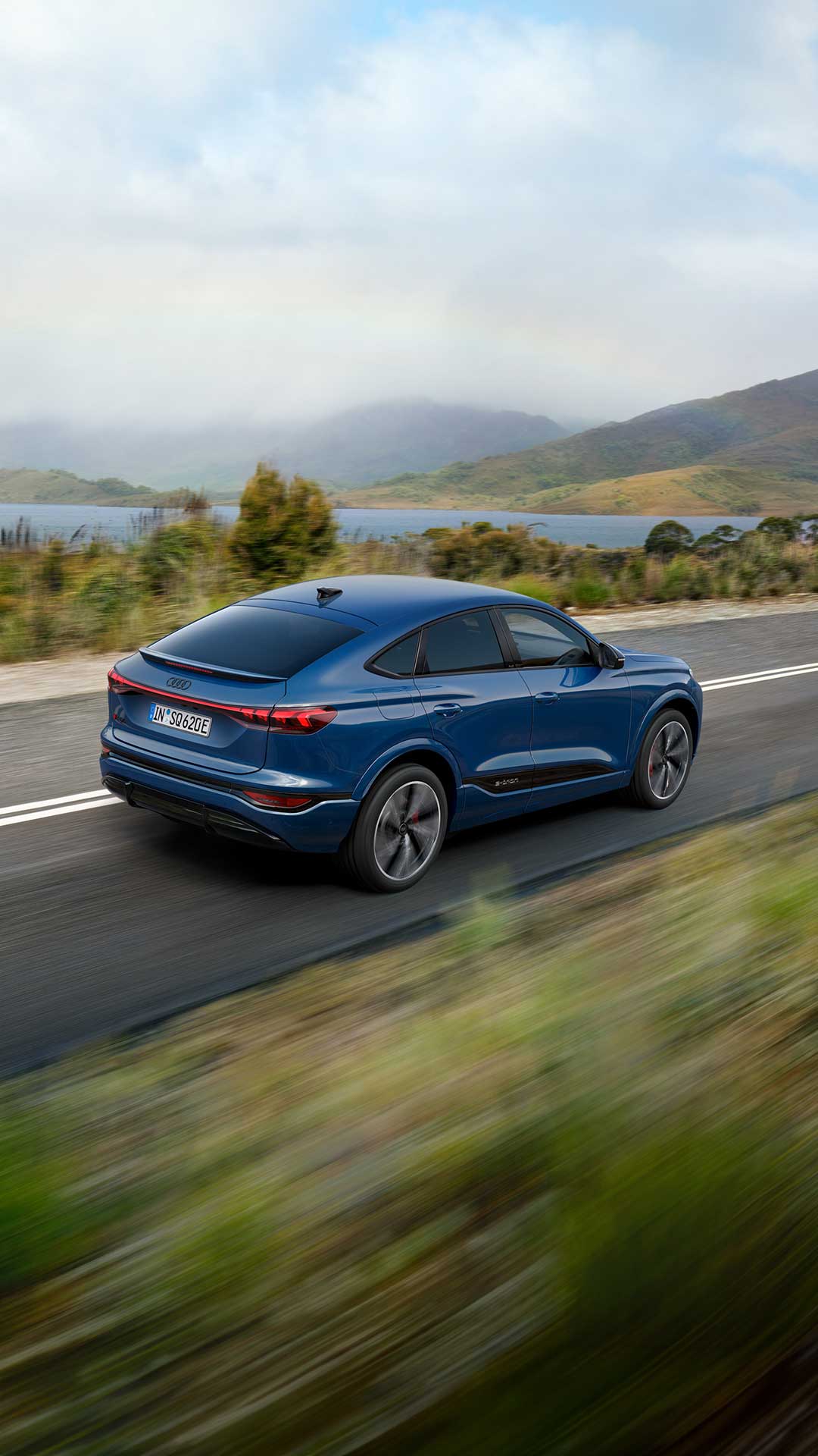 Ein blauer Audi SQ6 Sportback e-tron fährt auf einer Landstraße an See und Hügeln vorbei bei bewölktem Himmel.