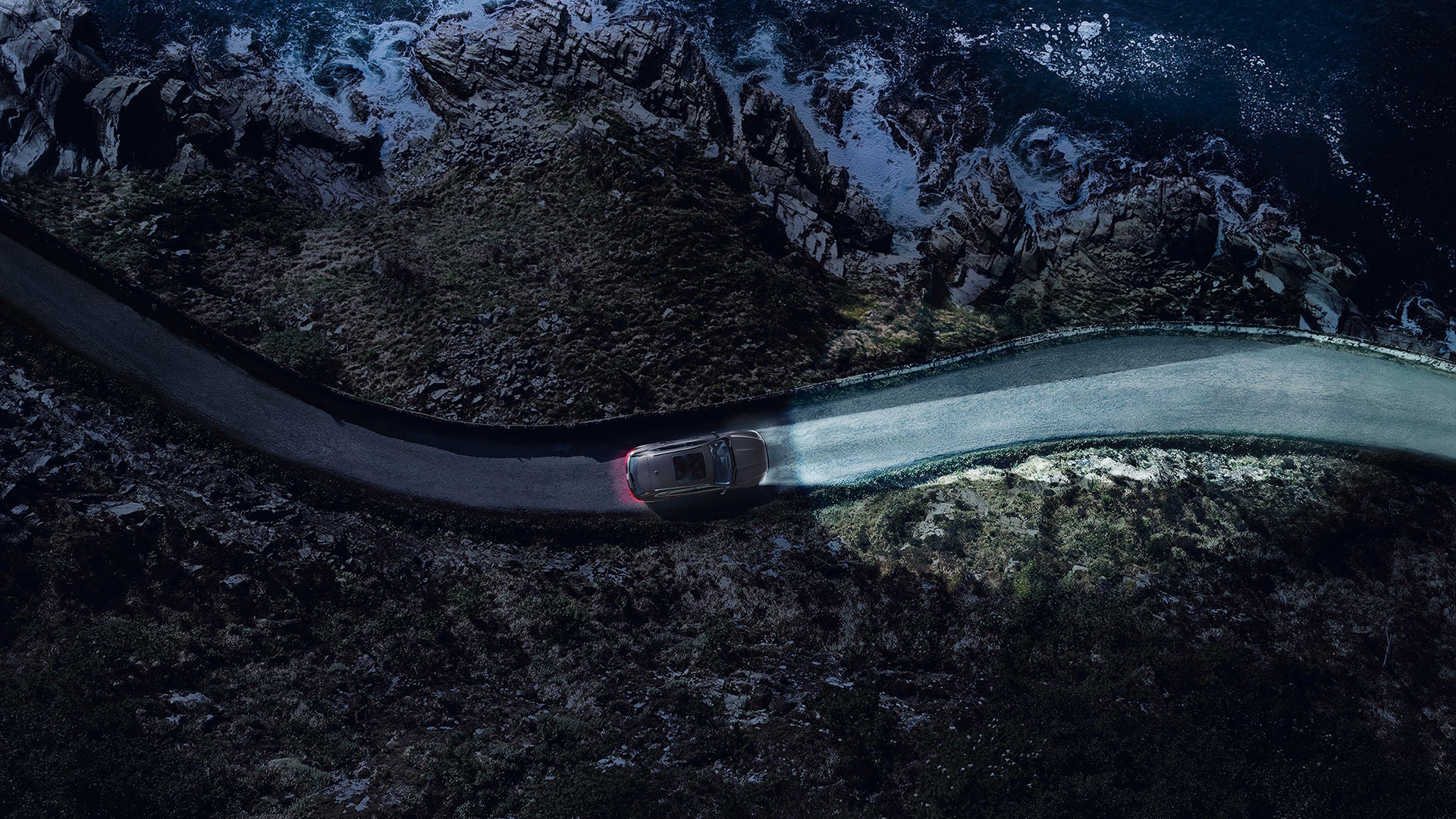 Auto mit eingeschaltetem Licht fährt bei Dunkelheit auf einer Küstenstraße entlang steiler Felsen und Wellen.
