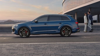 Blauer Audi Q7 SUV seitlich vor moderner Fassade, Paar im Businesslook steht lächelnd im Hintergrund.
