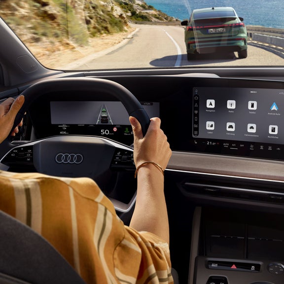Blick von hinten auf das Cockpit des Q4 SUV e-tron. Eine Frau sitzt am Lenkrad und steuert das Auto auf einer Bergstraße, zur rechten Wasser. Ein Fahrzeug fährt unmittelbar vor dem Q4 SUV e-tron. 