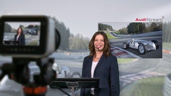 Woman with a headset stands behind a camera and is filmed in front of a racetrack screen.