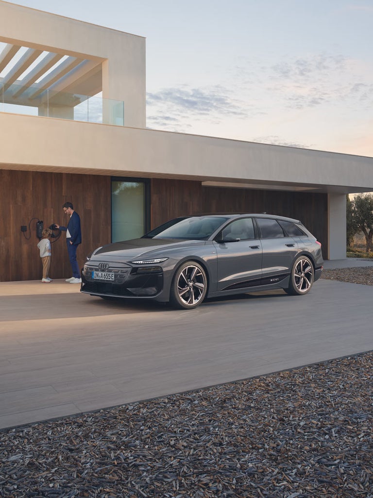 Grauer Audi A6 Avant e-tron parkt vor Haus, Vater lädt das Auto mit Kind an Wallbox.