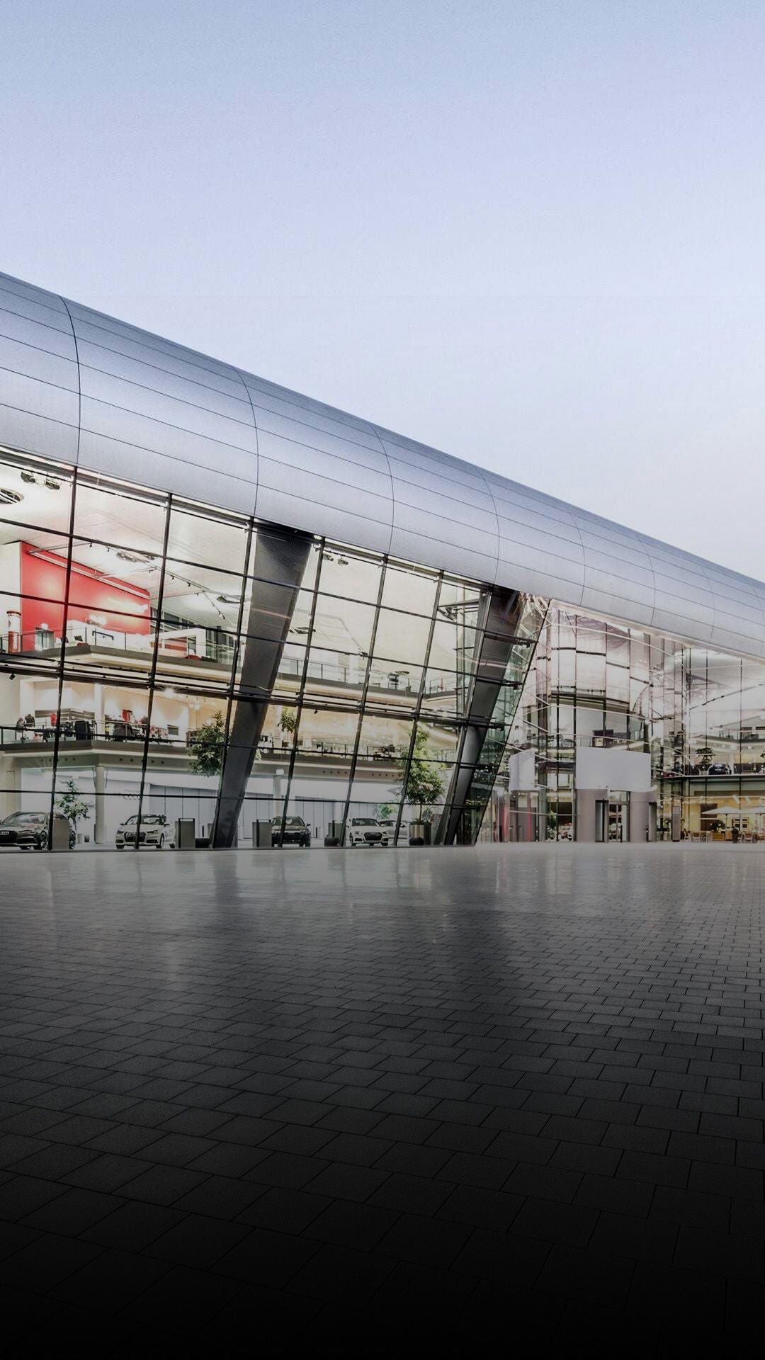 Moderne Glas- und Stahlfassade des Audi Forum Neckarsulm mit großzügigem Vorplatz und Audi-Logo bei Dämmerung.