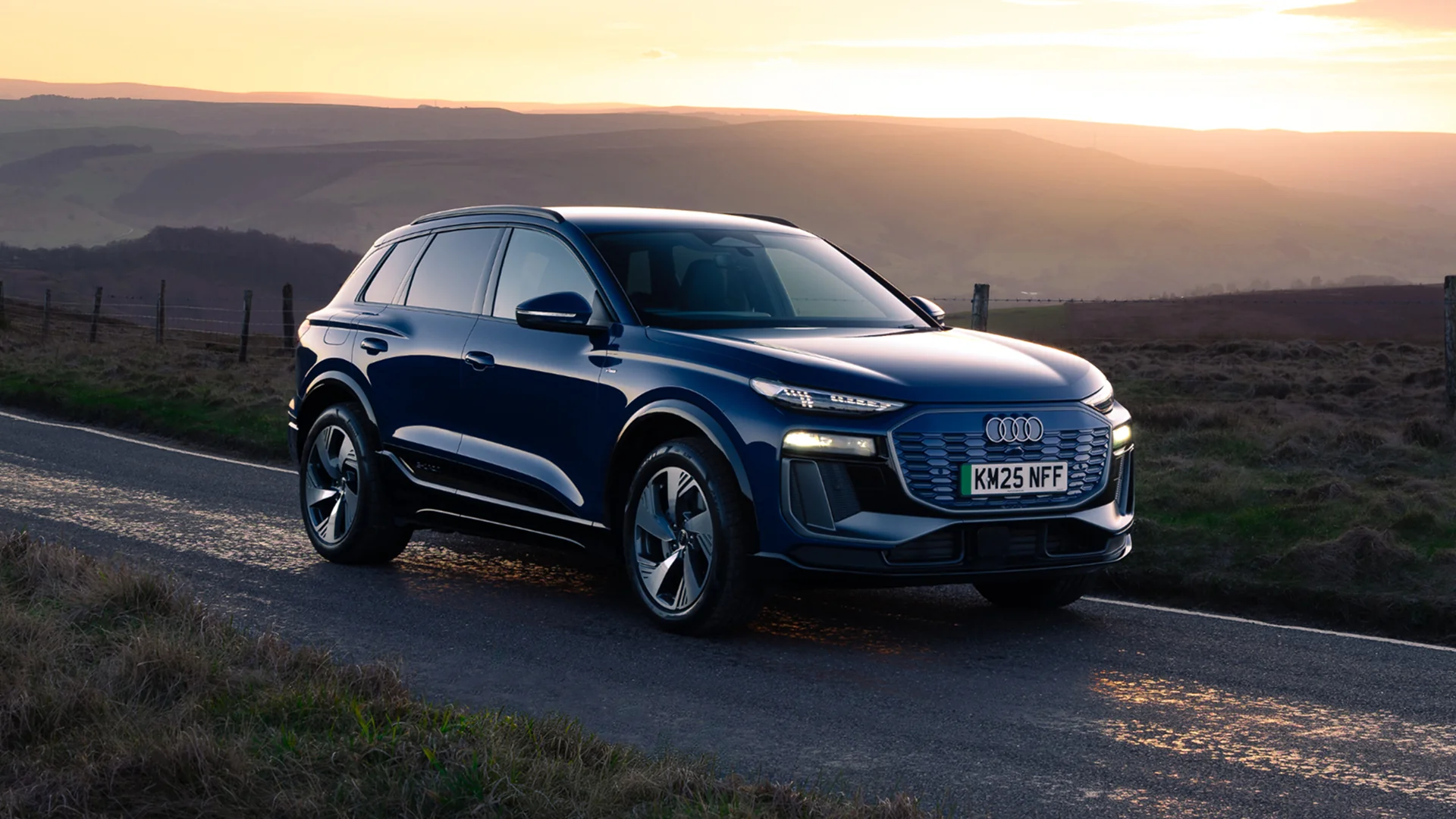 Blue Audi Q6 e-tron SUV parked on a rural road at sunset, shown from the front three-quarter angle. The vehicle features a wide illuminated grille, LED headlights and aerodynamic alloy wheels. Rolling hills and moorland stretch into the distance beneath a golden evening sky, with sunlight reflecting off the wet road surface.
