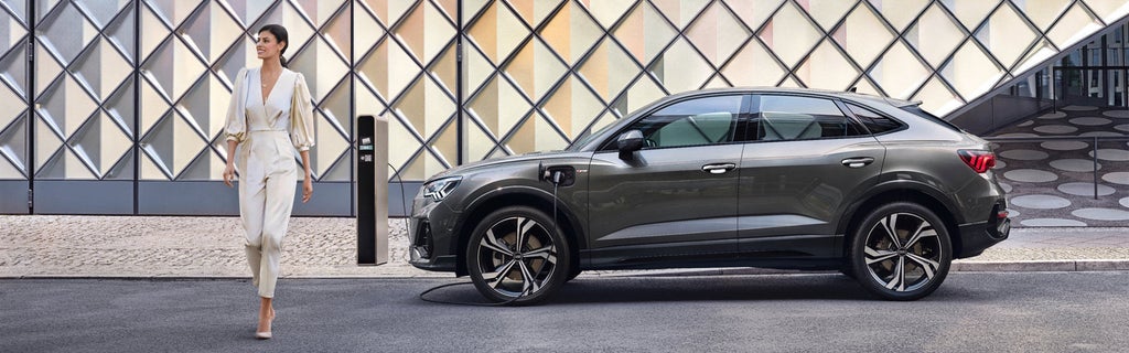  A grey Audi vehicle parked & on charge in an urban setting, a young woman walking away to the left