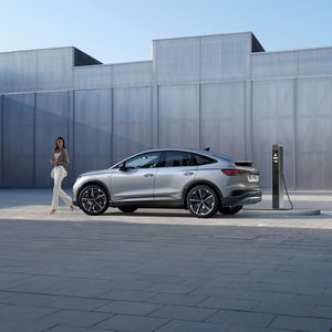A Audi SUV is parked at a charging station in front of a modern, minimalist building. A person stands nearby, conveying a sense of calm efficiency.
