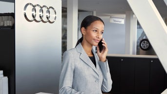 Frau im grauen Blazer telefoniert im Audi-Autohaus, neben ihr ein Schild mit Audi-Ringen.