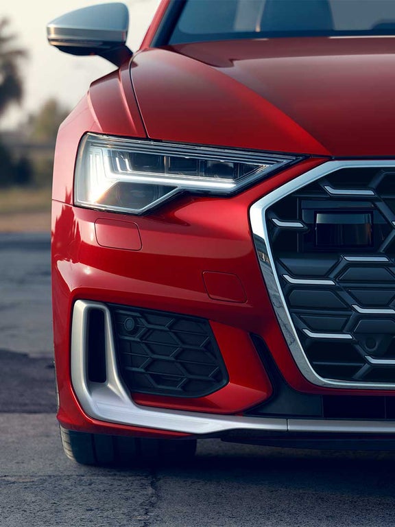 Nahaufnahme der Fahrzeugfront einer roten Audi S6 Limousine TDI mit Audi-Ringen und S-Logo im Kühlergrill, linkes LED-Frontlicht aktiv.