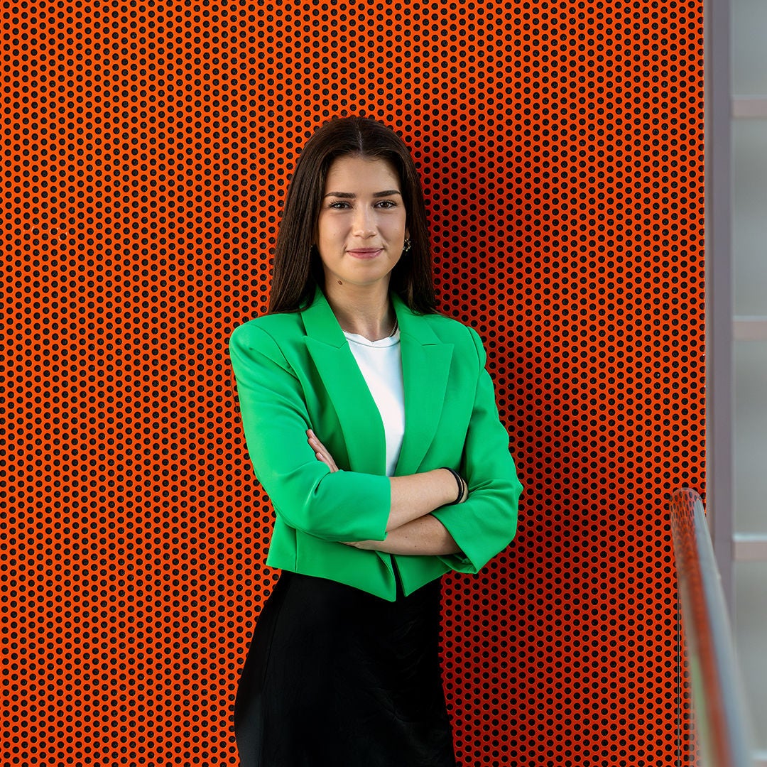Alina steht mit verschränkten Armen vor einer Wand und lächelt in die Kamera