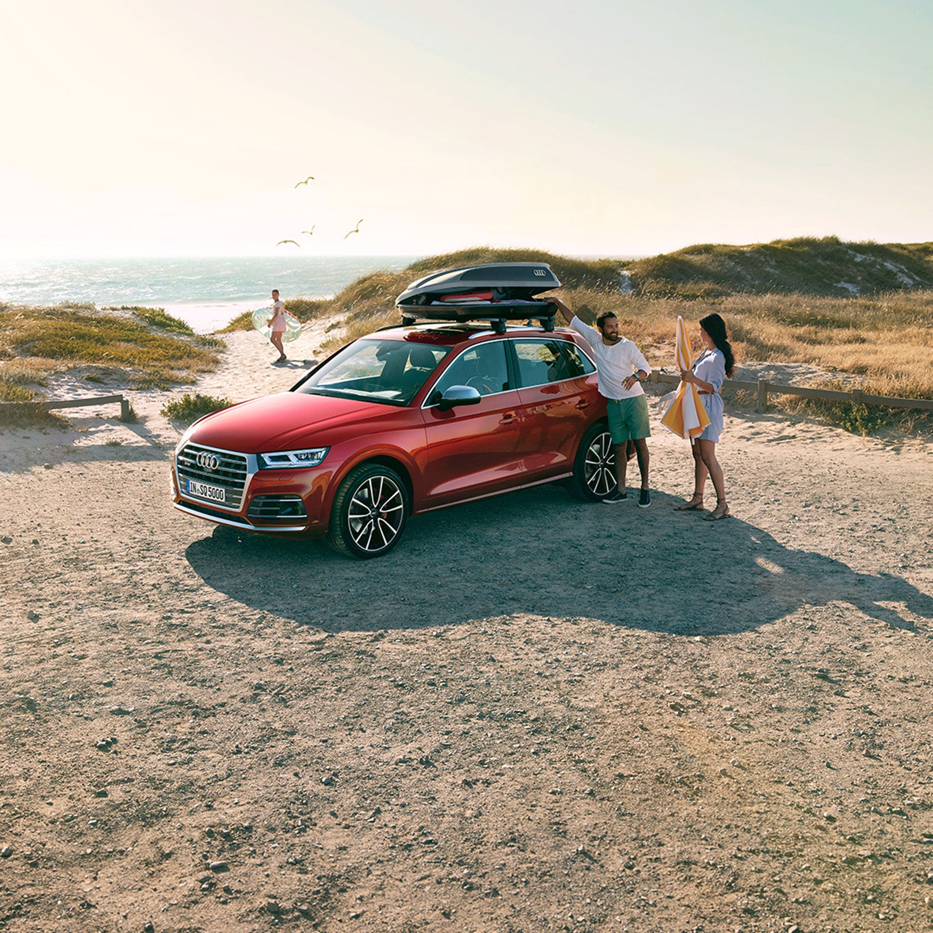 Ein roter Audi-SUV mit Dachbox steht am Strand, ein Paar packt aus, im Hintergrund läuft eine Person mit Surfbrett zum Meer.