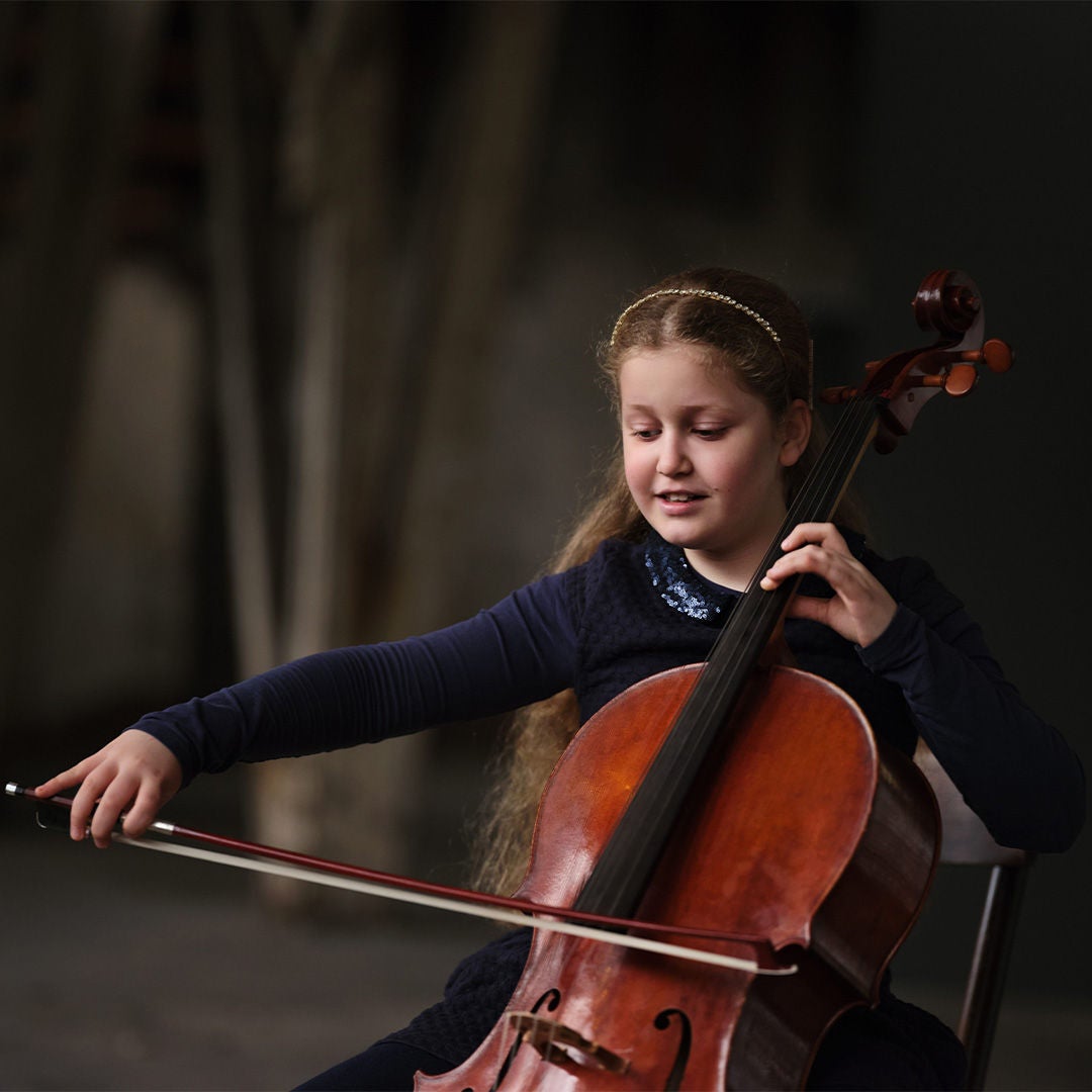 Junges Mädchen in Blau spielt konzentriert Cello vor dunklem, unscharfem Hintergrund.