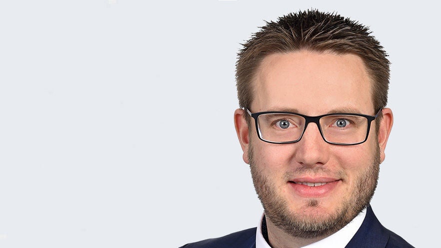 Magnus Weiß: Porträt lächelnder Mann mit Kurzhaarschnitt, leichtem Baat und Brille vor grauem Hintergrund.