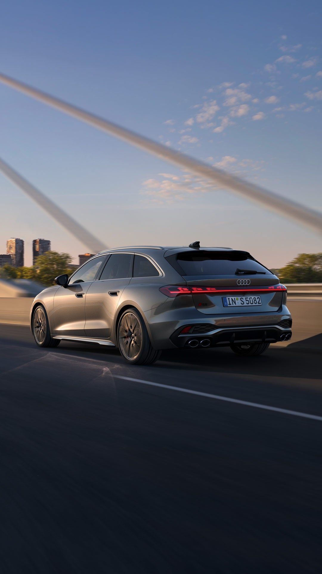 Grauer Audi S5 Avant fährt über eine Brücke mit Stadt-Skyline im Hintergrund bei Sonnenuntergang.