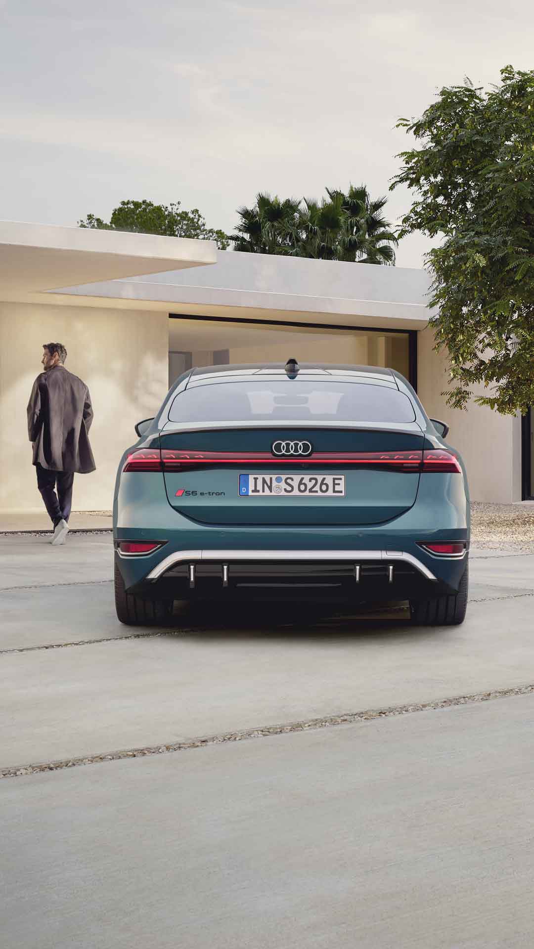 Der S6 Sportback e-tron im Heckprofil. Das Fahrzeug ist dem Eingangsbereich eines modernen Gebäudes zugewandt. Vor dem Auto befindet sich ein junger Mann, der auf das Gebäude zuläuft.