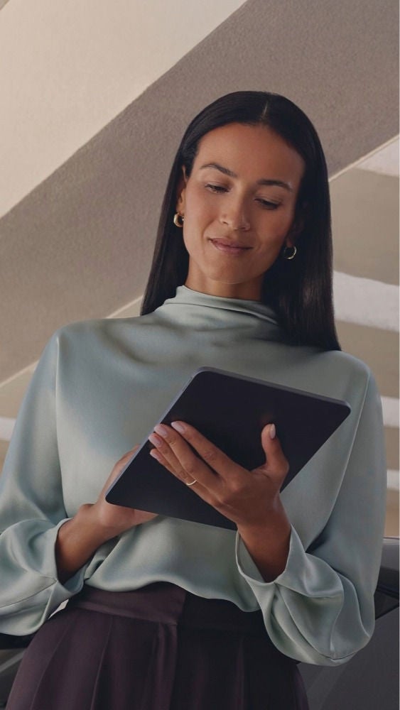 A person holding a tablet stands in front of a car, under a modern, sunlit covered structure.