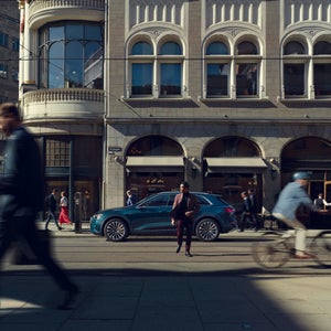 A man stands confidently on a bustling city street beside a blue SUV, surrounded by blurred motion of pedestrians and a cyclist, against historic architecture.