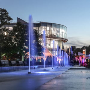 Moderne Glasfassade des Audi Forums bei Dämmerung, bunte Lichtfontänen und Besuchende im Vordergrund.