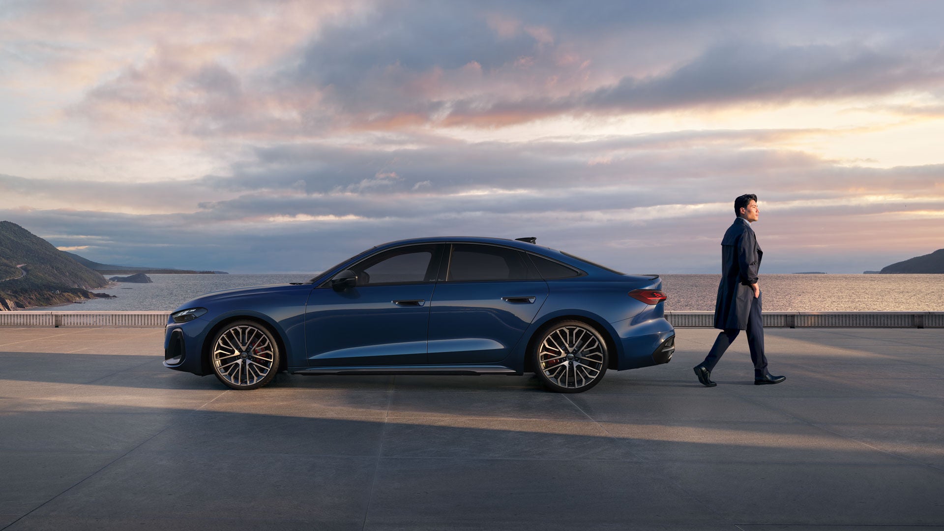 Blaue Audi S5 Limousine steht auf einer Küstenplattform bei Sonnenuntergang, ein Mann im Mantel geht daran vorbei in Richtung Meer.