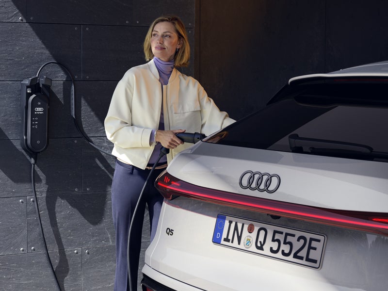 Blick auf das Heck des Q5 Sportback e-hybrid. Eine junge Frau schließt das Fahrzeug an eine Ladestation an.