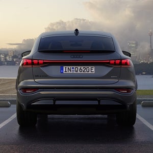 Heckansicht des Q6 Sportback e-tron, welcher auf einem Parkplatz am Meer steht. Im Hintergrund sieht man eine Stadt, welche von Wolken durchzogen ist.