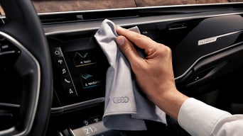 Hand wischt Touchscreen im Auto mit einem Mikrofasertuch mit Audi-Logo.
