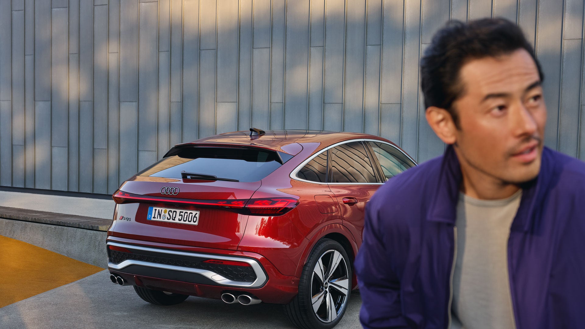 Ein roter Audi SQ5 Sportback steht vor einer grauen Wand, im Vordergrund blickt ein Mann im lila Jackett zur Seite.