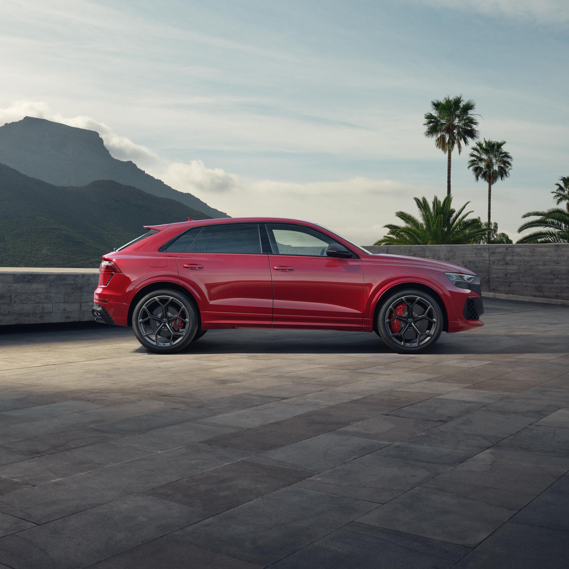 Seitenansicht eines roten Audi RS Q8 SUV performance auf heller Steinfläche vor Berglandschaft und Palmen bei Tageslicht.