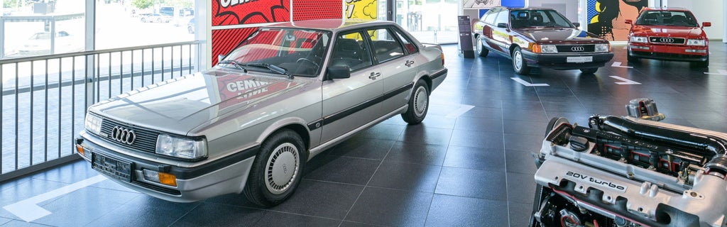 An exhibition featuring several historic Audi vehicles, an engine, and colorful comic book posters.