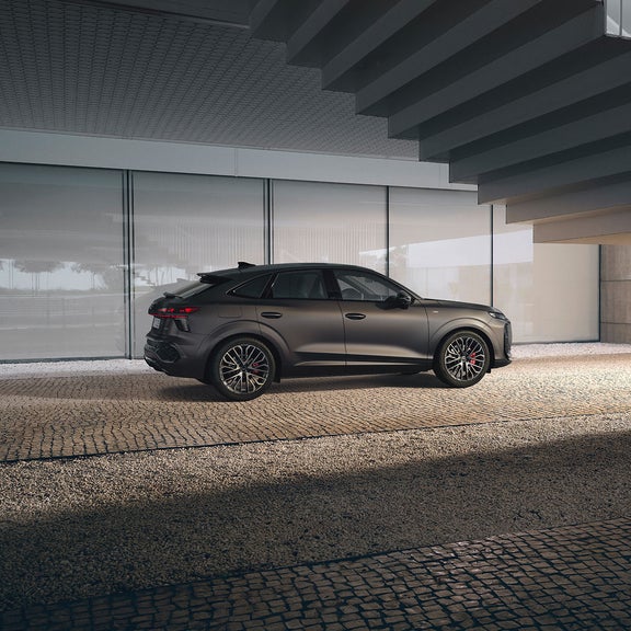 A matte black Audi Q3 parked in a modern covered area with glass walls and cobblestone ground.