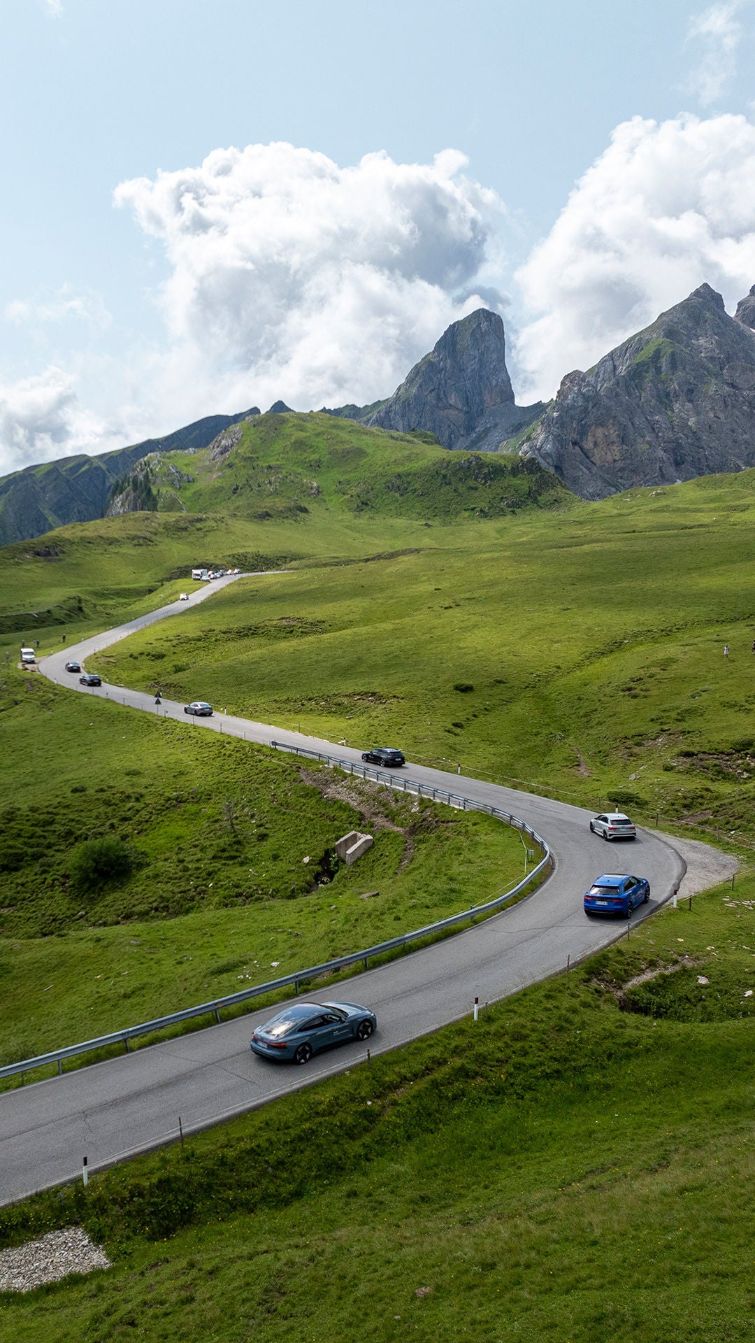Bird's eye view of a landscape with a winding road on which several Audi models are driving one behind the other, with mountains in the background.