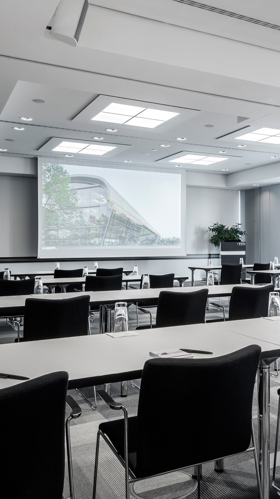 Leerstehender Konferenzraum mit Reihen aus Tischen, Stühlen, Block und Glas, Blick zur Leinwand.