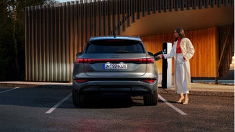 A woman in stylish attire plugs in an Audi SQ6 e-tron to a charging station, set against a sleek wooden structure.