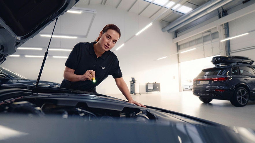 Mitarbeiterin prüft Motorraum eines dunklen Audi in heller Werkstatt mit Taschenlampe.