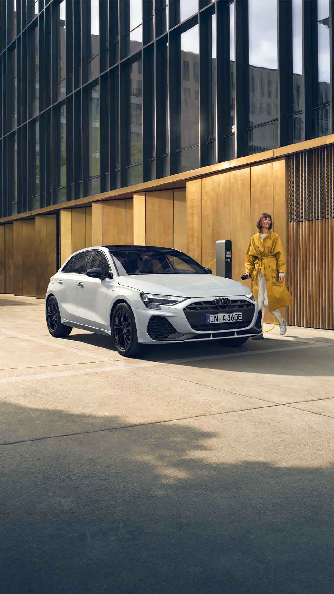 Weißer Audi A3 Sportback TFSI e steht an Ladesäule, Frau im gelben Mantel mit Kamera lächelt und geht daran vorbei.