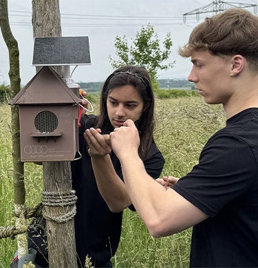 Two young people install a solar-powered listening station in the form of a nesting box in a meadow.