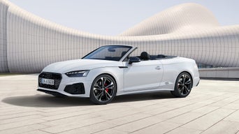 Weißes Audi A5 Cabriolet mit geöffnetem Verdeck steht vor moderner, geschwungener Gebäudefassade auf heller Steinfläche.