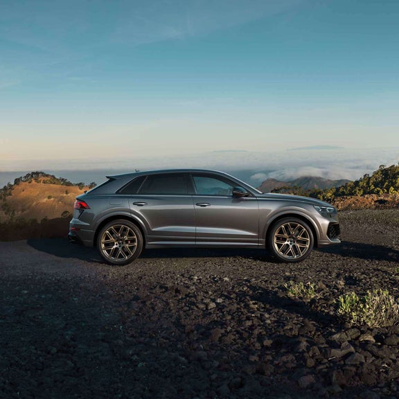 Seitenansicht eines grauen Audi RS Q8 SUV vor bergiger Landschaft mit Bäumen und Wolken im Hintergrund.