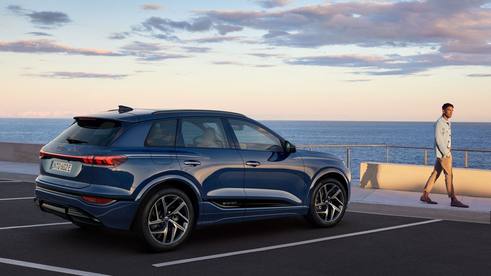 Blauer Audi-SUV steht auf einem Parkplatz am Meer, ein Mann in heller Kleidung geht am Geländer entlang.