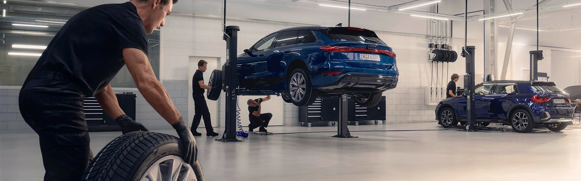 Technicians servicing Audi vehicles in a workshop, with one vehicle raised on a lift and maintenance work being carried out.